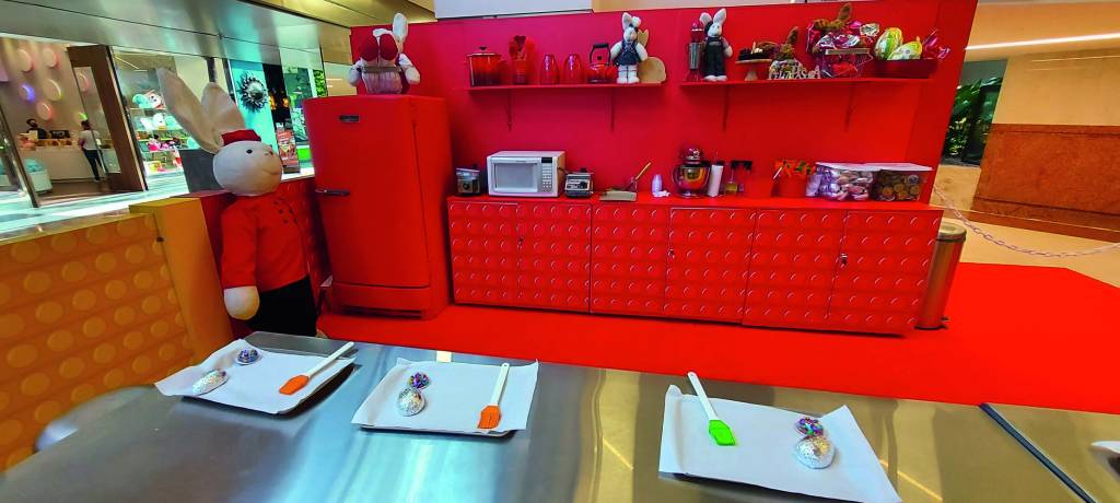 PODE SER ESTA divulgação (1).jpg Uma cozinha temática do brinquedo pop it, com geladeira, armários e demais utensílios vermelhos. No centro, um grande balcão tem bandejas e espátulas para a confecção de ovos de Páscoa