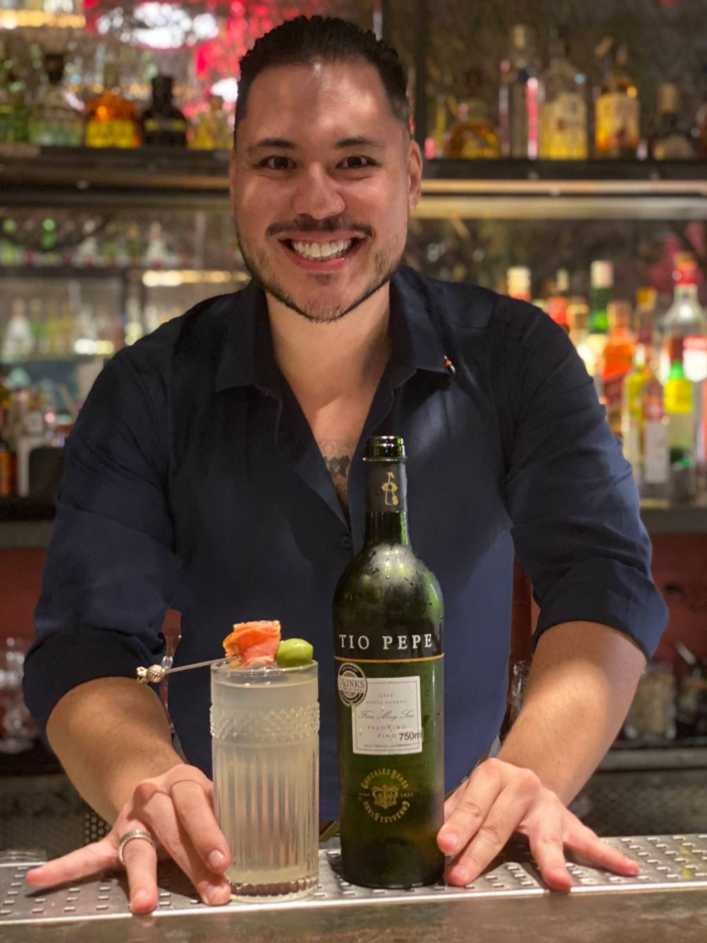 Tio Pepe Challenge Brasil 2022 Ricardo Takahashi Paulon posa em balcão de bar junto de drinque de cor clara servido em copo alto guarnecido de presunto e azeitona verde