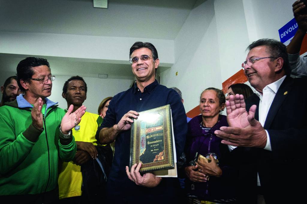 No Bom Prato Paraisópolis, Rodrigo Garcia é empossado novo gov Imagem mostra homem sorrindo em volta de pessoas que aplaudem. Ele sorri.