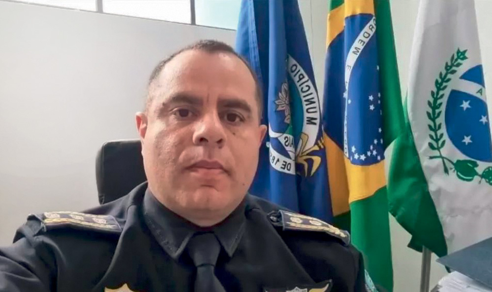 SECRETÁRIO.JPG Imagem mostra homem vestindo farda escura sentado em frente a duas bandeiras.