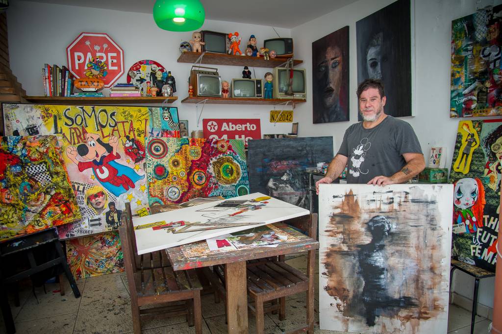 Vímerson Cavanilas (Rogério Pallatta) Vímerson Cavanilas posa em área de trabalho com prateleiras repletas de televisões analógicas, bonecos antigos, peças coloridas garimpadas por ele e os quadros. Ele posa de pé, atrás de um quadro com tons neutros, e veste camiseta cinza.
