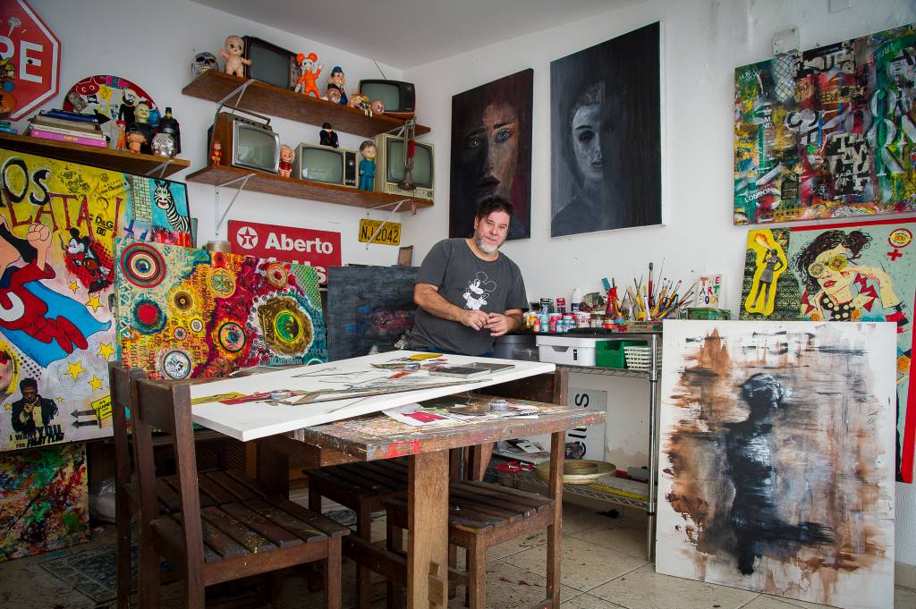 Vímerson Cavanilas (Rogério Pallatta) Vímerson Cavanilas posa em área de trabalho com prateleiras repletas de televisões analógicas, bonecos antigos, peças coloridas garimpadas por ele e os quadros. Ele posa sentado e veste camiseta cinza e jeans escura, em frente à mesa de trabalho.