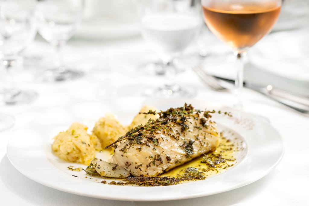 Bistrot Parigi Em um prato raso de louça branca, é servido um lombo de bacalhau assado junto de legumes e molho de manjericão