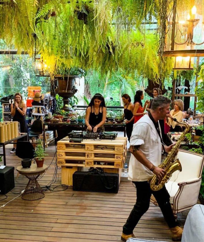 Bazar Mamma Mia Foto de um espaço aberto, com mesas, estandes e plantas penduradas no teto, onde acontece um bazar. Um homem toca saxofone e uma DJ toca em um estande de madeira