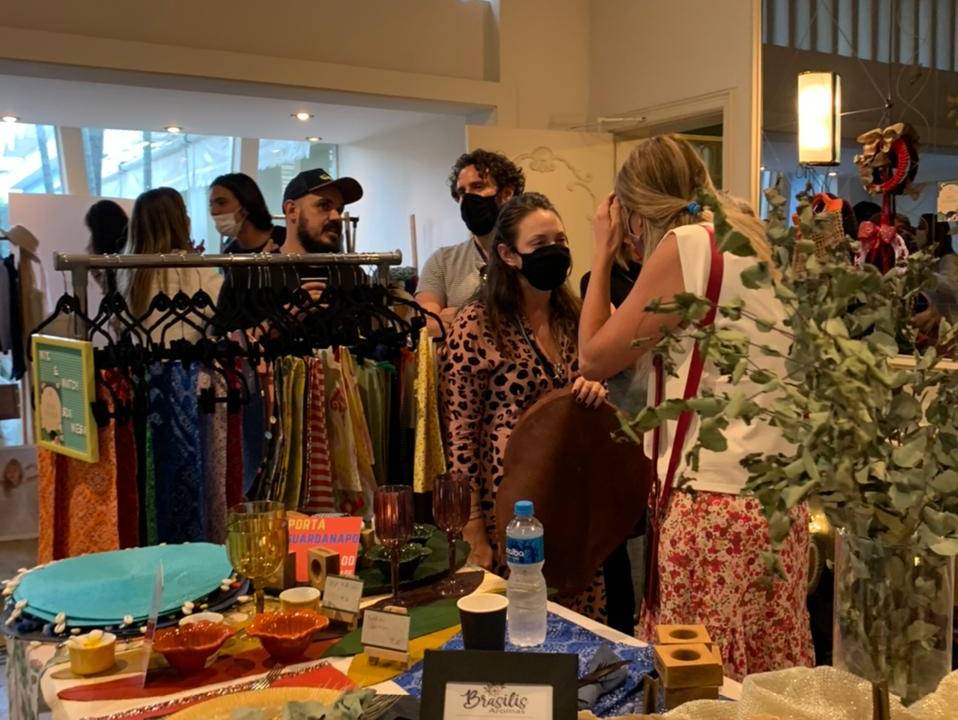 Pessoas compram em um bazar, onde se vê araras de roupas e mesas com produtos diversos