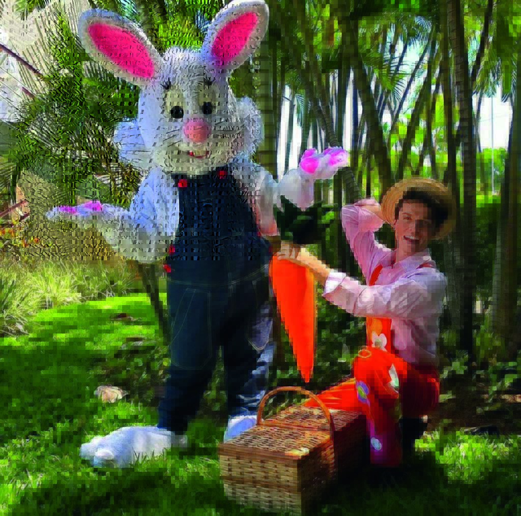 divulgação.jpg Um ator fantasiado ao lado de outro ator fantasiado de coelho da Páscoa posam para uma foto em um jardim com grama. O ator, ajudante do coelho, segura uma cenoura gigante de mentira e uma cesta de piquenique