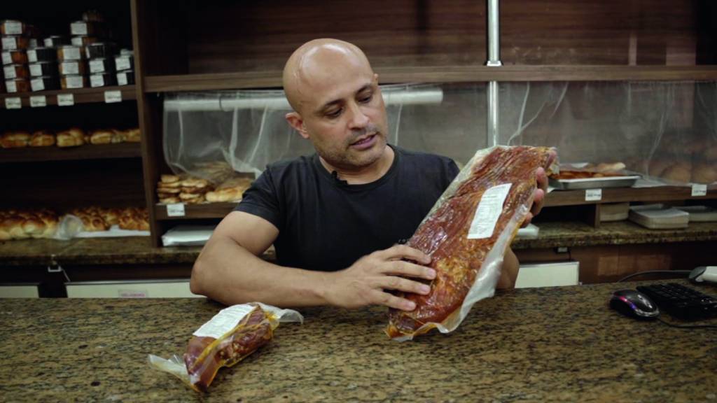 emporium-lado-a-lado-1.jpg Homem branco, magro e careca sentado em uma mesa. Ele segura pacotes de carne de seu estabelecimento
