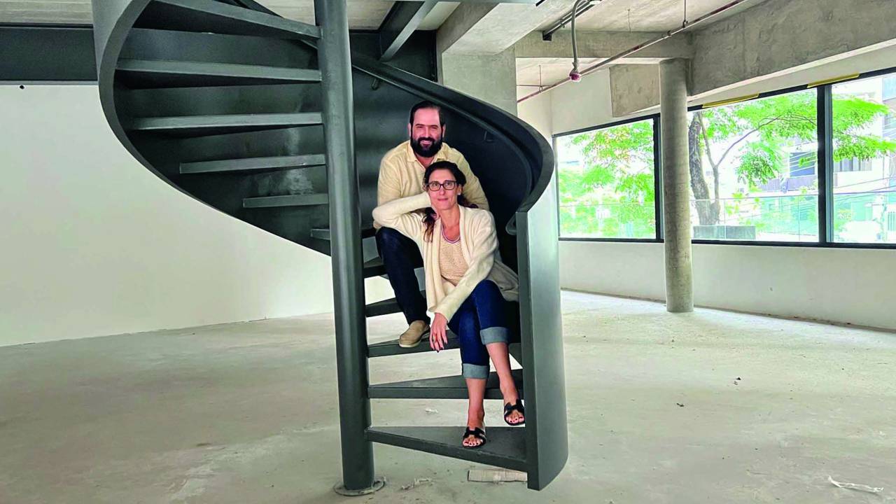 Imagem mostra um homem e uma mulher sentados em uma cadeira caracol preta, no centro da imagem. Ao redor, um espaço vazio, de concreto.