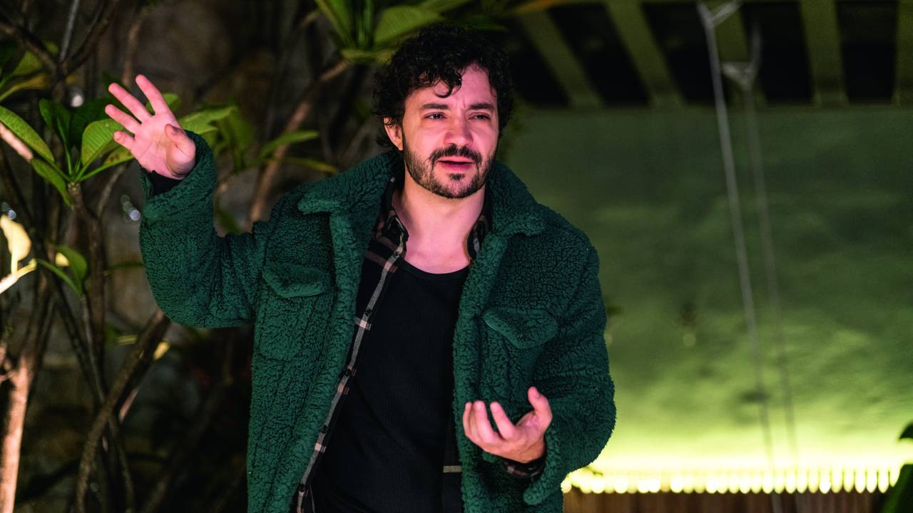 Rafael Primot é um homem branco e magro, de cabelos e barbas escuras. Ele usa uma camiseta preta e uma jaquete verde escura e gesticula com uma das mãos para o alto