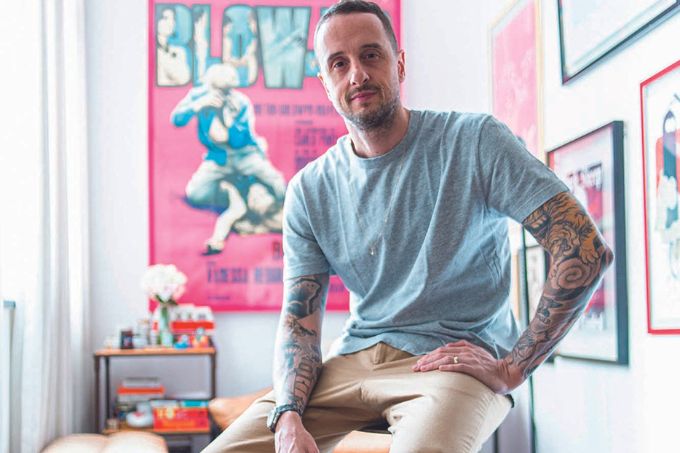 Hector Miranda, um homem branco, de cabelos e barba escura, posa para a foto sentado em um banco em uma sala com carpete colorido e quadros coloridos nas paredes