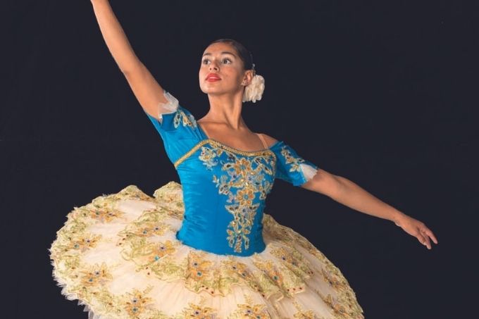 Bailarina negra, de coque, em posição da dança. Ela usa um figurino azul com tutu dourado