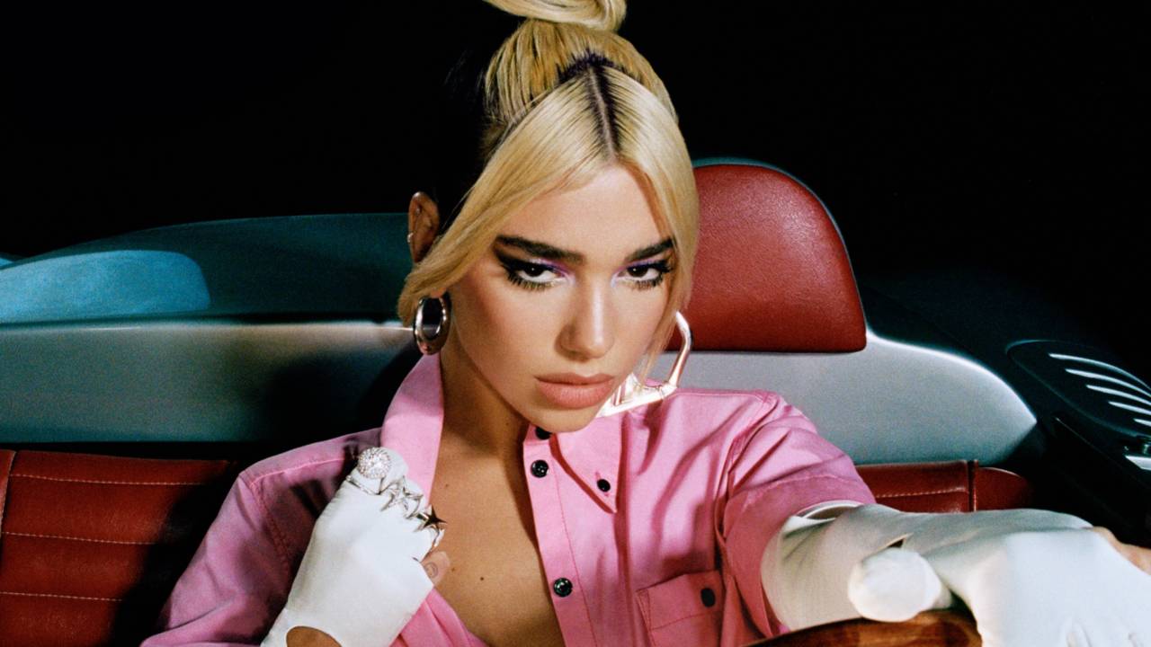 Imagem mostra mulher dentro de carro, com uma mão no volante. Ela tem cabelo loiro e veste roupa rosa com mangas brancas