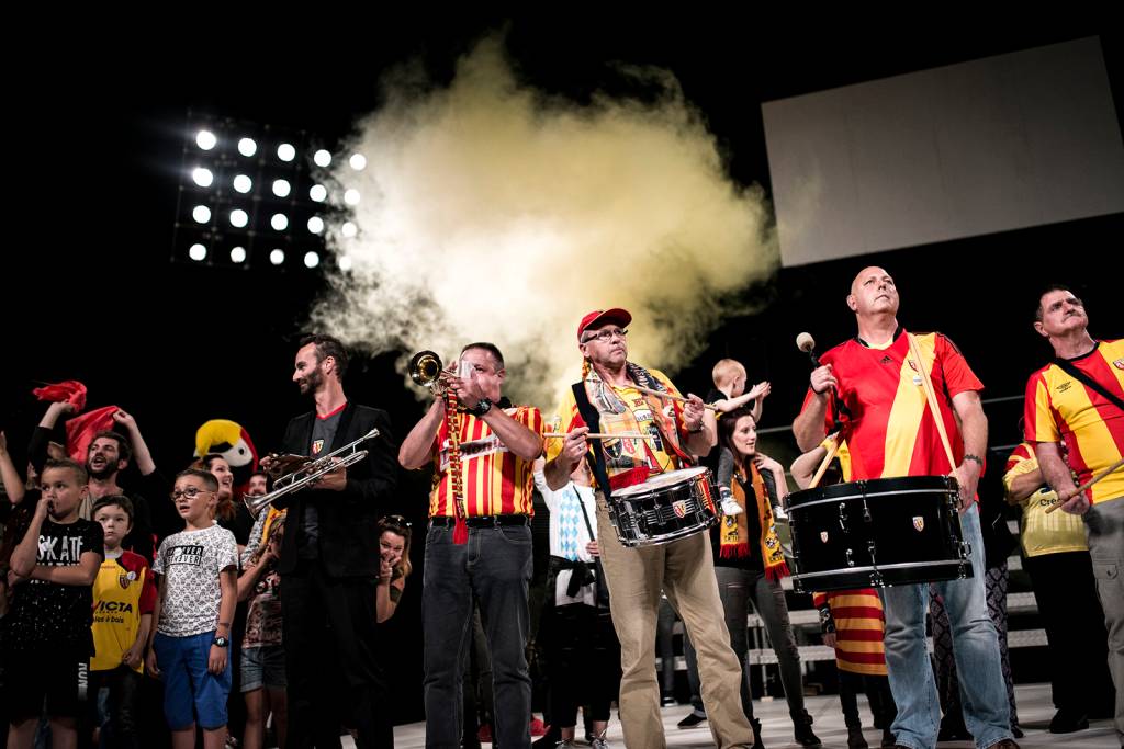 STADIUM_©Yohanne Lamoulère – Tendance floue (2) Torcedores tocam instrumentos em grupo no palco. Usam camisetas vermelhas e amarelas