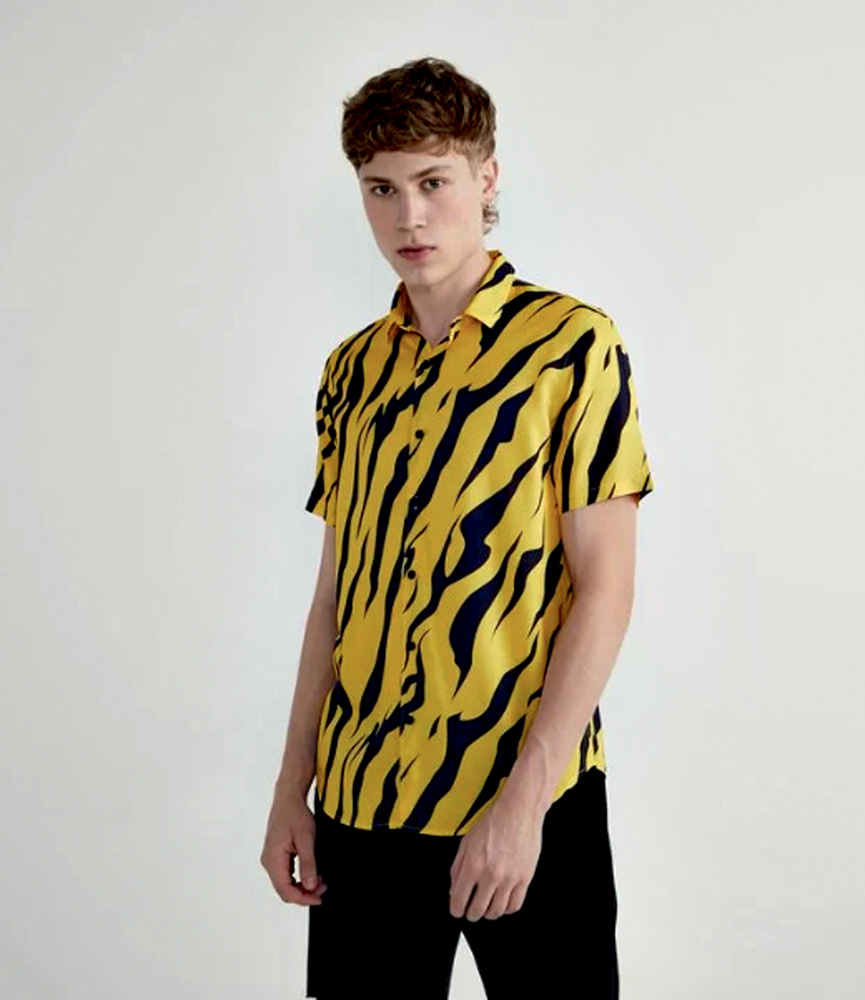 Modelo branco, magro e de cabelos curtos e castanhos veste camisa de maga curta amarela com estampa de tigre