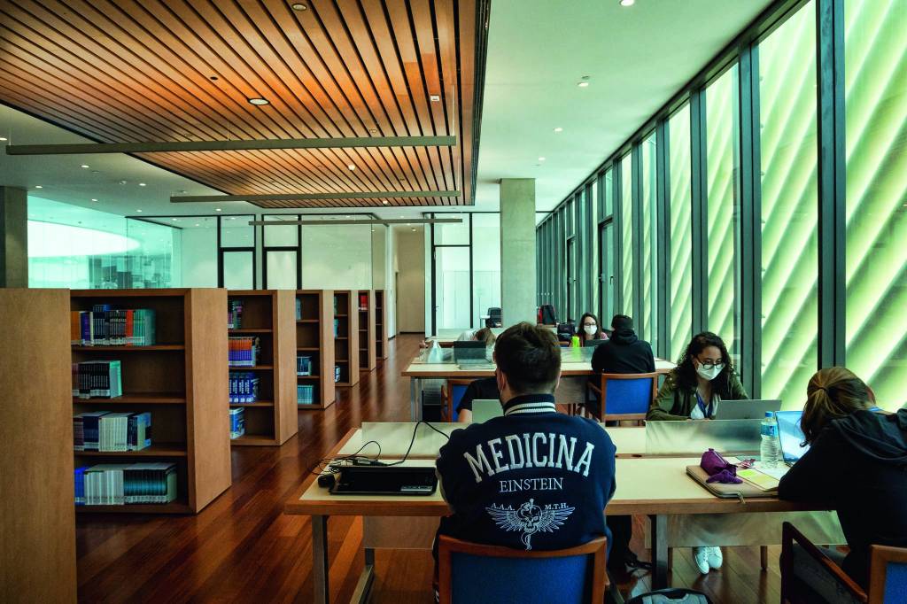 Sidney Klajner Alunos estudam sentados em mesas da biblioteca. O ambiente, com estantes, é aconchegante