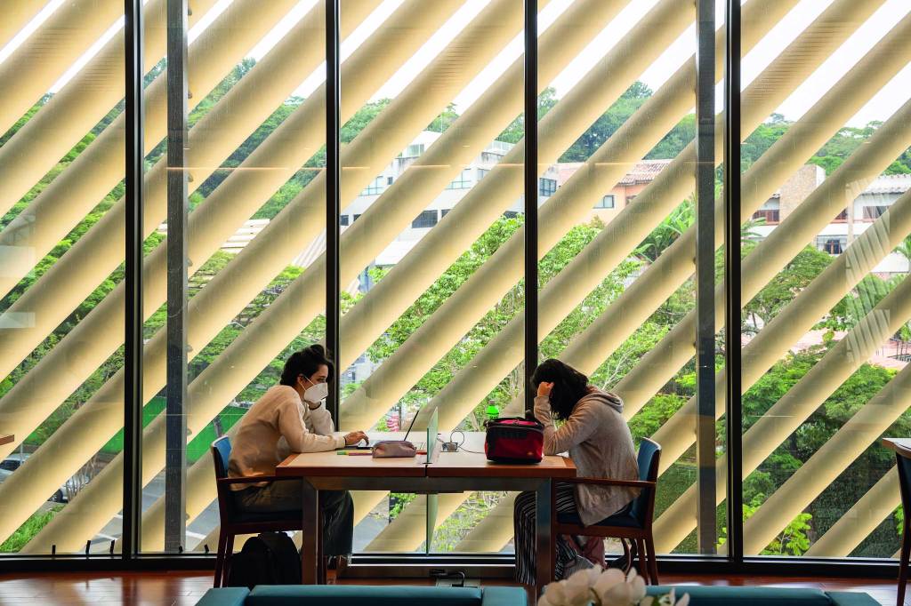Sidney Klajner Duas estudantes sentadas em uma mesa de uma das cafeterias do prédio. A parede permite que frestas de luz passem, o que deixa o ambiente aconchegante