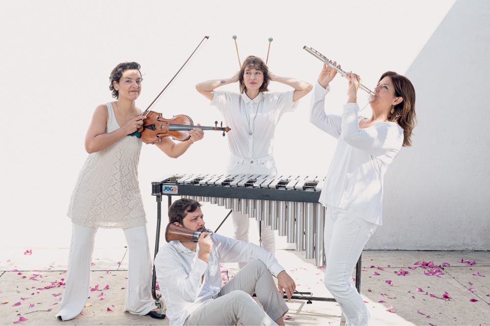 Quarteto de músicos, com três mulheres e um homem, posa para foto. Todos usam roupas brancas e estão em um fundo branco. Uma das três músicas, que estão todas em pé, toca um violino, a do meio toca um instrumento de percussão similar a um xilofone, e a do canto direito toca uma flauta. O homem, no centro, está sentado no chão e toca um instrumento de sopro marrom