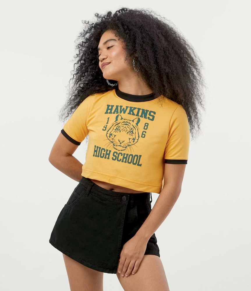 Cropped.jpg Modelo negra, alta e magra, de cabelos crespos compridos, usa cropped amarelo com a estampa de Hawkins High