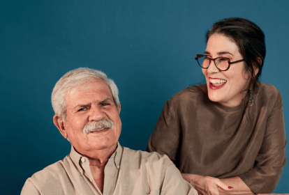 Imagem mostra homem de cabelo e bigode grisalho sorrindo ao lado de mulher de óculos e roupa marrom, sorrindo também