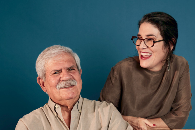 Imagem mostra homem de cabelo e bigode grisalho sorrindo ao lado de mulher de óculos e roupa marrom, sorrindo também