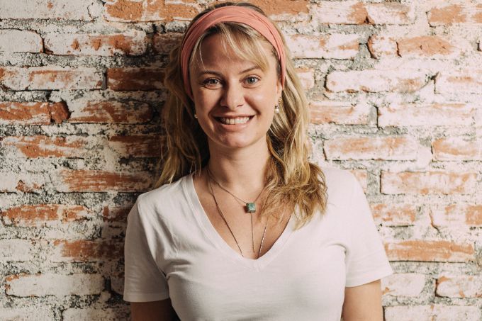 Luiza é uma mulher branca, magra, loira e jovem. Usa camiseta branca, tiara rosa e um avental de cozinha. Posa sorrindo em frente a uma parede de tijolos brancos