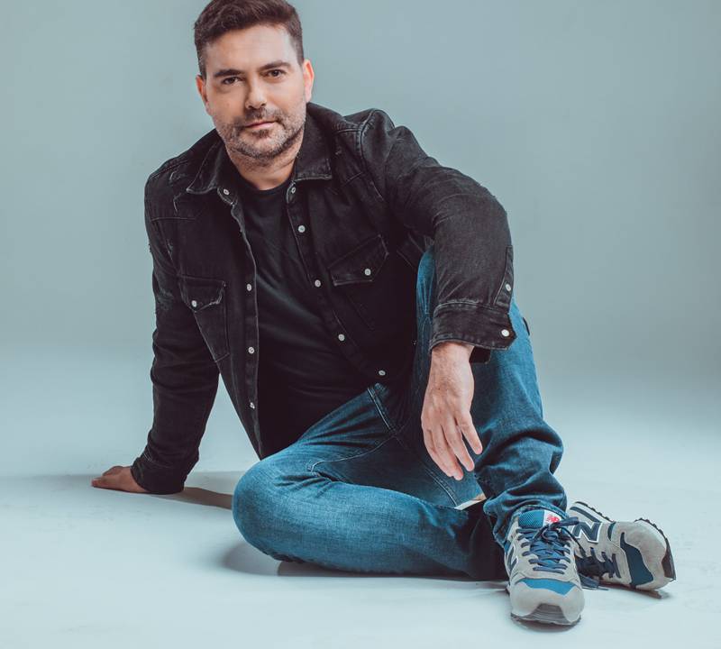 Rodrigo Toigo posa sentado no chão com um joelho dobrado. Veste jeans azul e jaqueta preta.