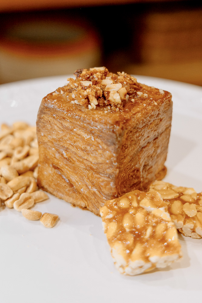 Zestzing Padaria Artesanal Doce em forma de cubo, coberto por pedaços de amendoim e rodeado por pedaços de pé-de-moleque sobre um prato raso de cor branca