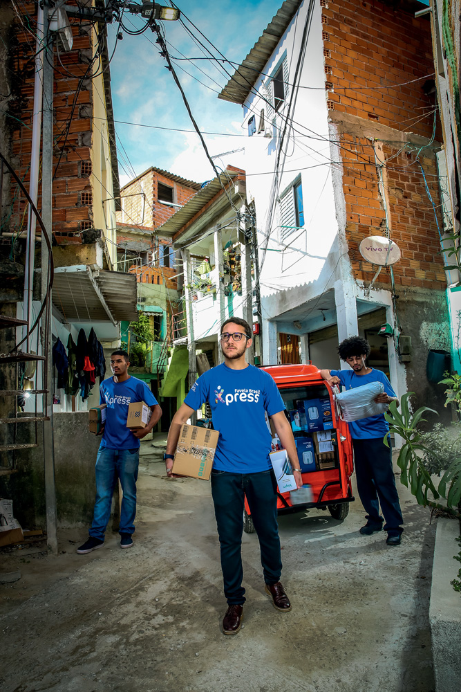 FAVELAEXPRESS_5.JPG Giva, um homem branco, de cabelos e barba castanhos, veste uniforme de sua empresa de entregas. Está na frente de outros dois trabalhadores, homens negros que também usam uniforme, em uma das ruas de Paraisópolis. Eles seguram pacotes e caixas e compras on-line