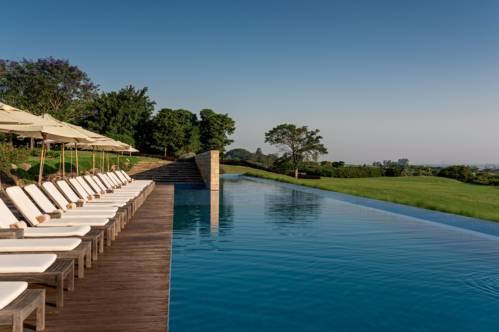 Foto exibe piscina de borda infinita com cadeiras de praia brancas à esquerda e chão de madeira. Ao fundo, árvores e gramado.