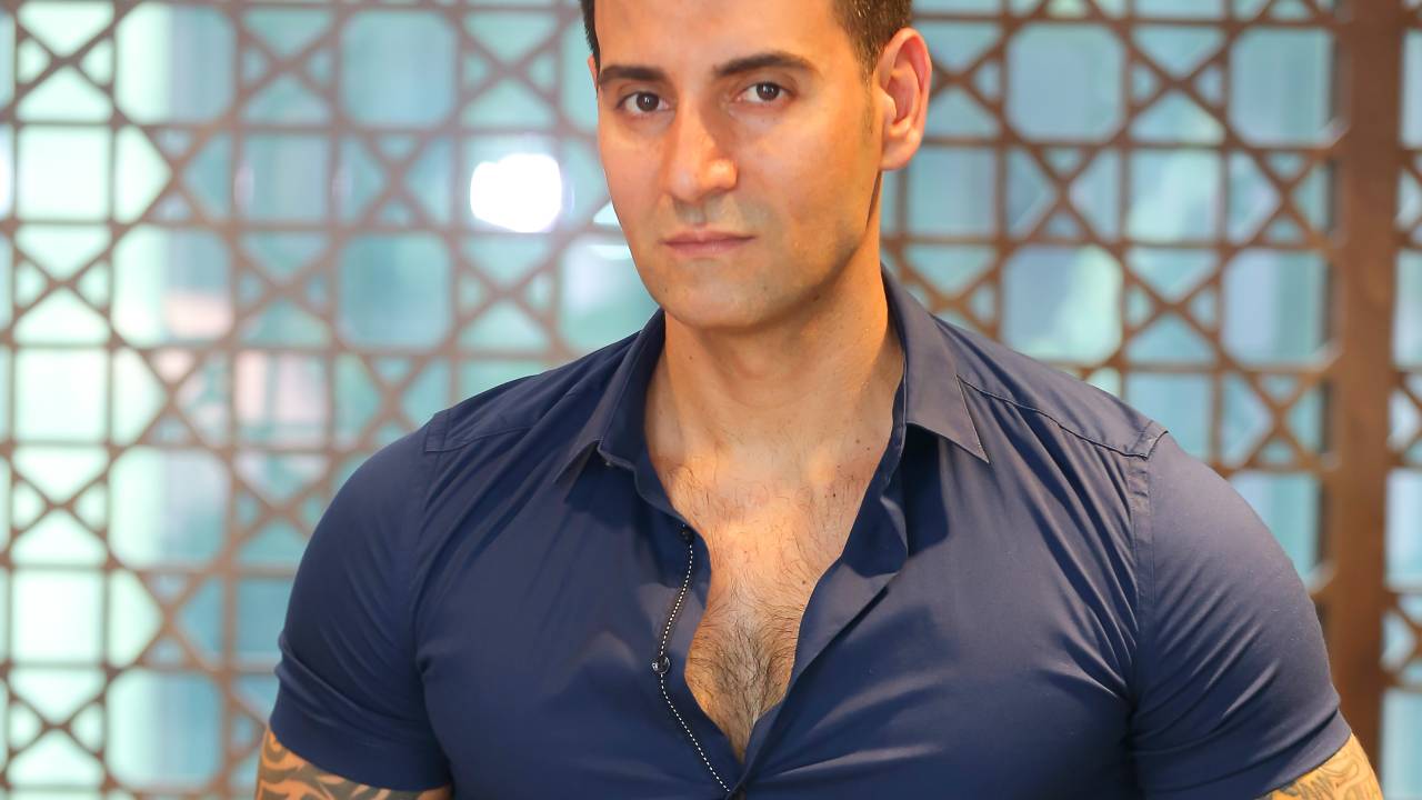Estilista e arquiteto Vincenzo Visciglia posa com uma mão no bolso da calça preta com cinto de couro. veste camisa justa de manga curta na cor azul-marinho. Nos braços aparecem tatuagens.