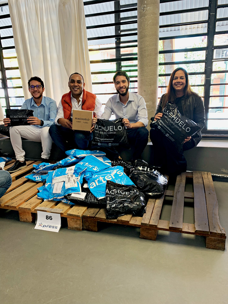 Riachuelo.jpeg Giva e seus funcionários e parceiros sentados com vários pacotes de encomendas em um suporte de madeira à frente