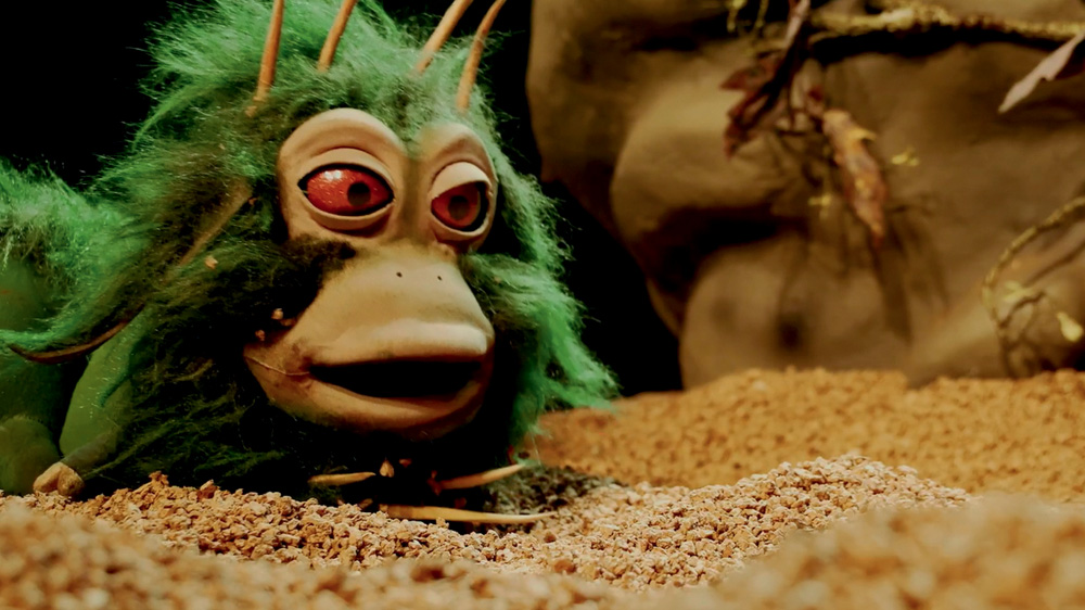 VIk espetáculo (1).jpg Boneco que parece um lagarta, com olhos vermelhos e pelo verde. Ele está em um palco com areia