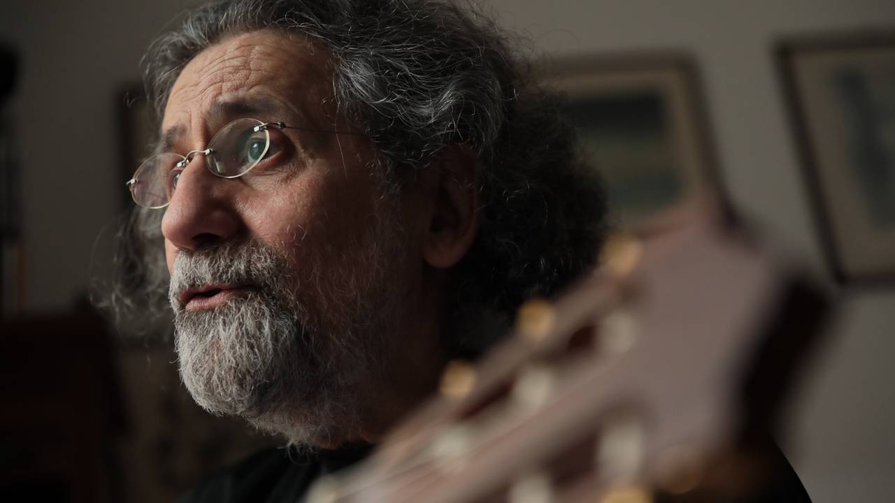 Imagem mostra homem de barba grisalha e óculos segurando violão