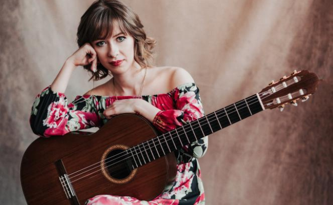 Imagem mostra mulher com violão no colo olhando para a câmera. Ela veste um vestido estampado, florido.