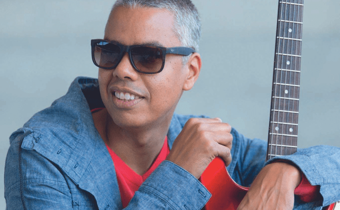 Imagem mostra homem grisalho com óculos escuros sorrindo e abraçando uma guitarra vermelha