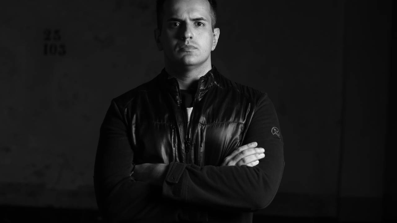 Luís Aizcombe, criador da Corbe Toys, posa com jaqueta de couro e braços cruzados. Aparece careca e com a cara séria em foto preto e branco.