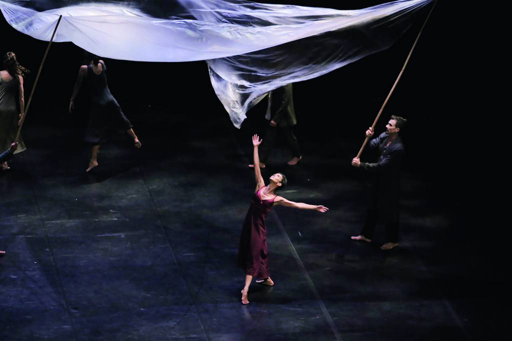PÁGINA 4 – Odisseia, São Paulo Cia. de Dança (RODOLFO DIAS PAES-DIVULGAÇÃO).jpg Dançarina no meio do palco, em movimento com os braços para cima. Usa vestido vinho reto e comprido e está rodeada por outros dançarinos que elevam panos brancos