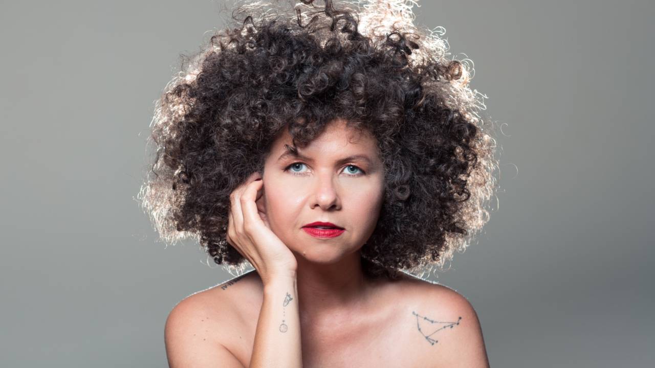 Imagem mostra mulher com uma das mãos sobre o rosto. O fundo é cinza.