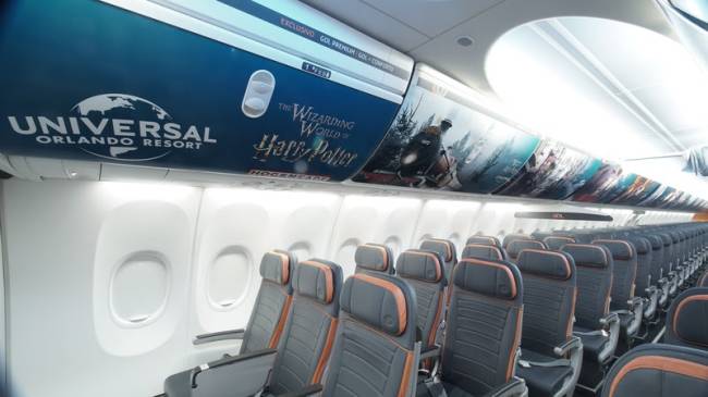Avião do Harry Potter Foto de interior de avião, com quatro fileiras de cadeiras com três assentos. Na parte de cima, bagageiro com adesivo que mostra letreiro e cena de Harry Potter