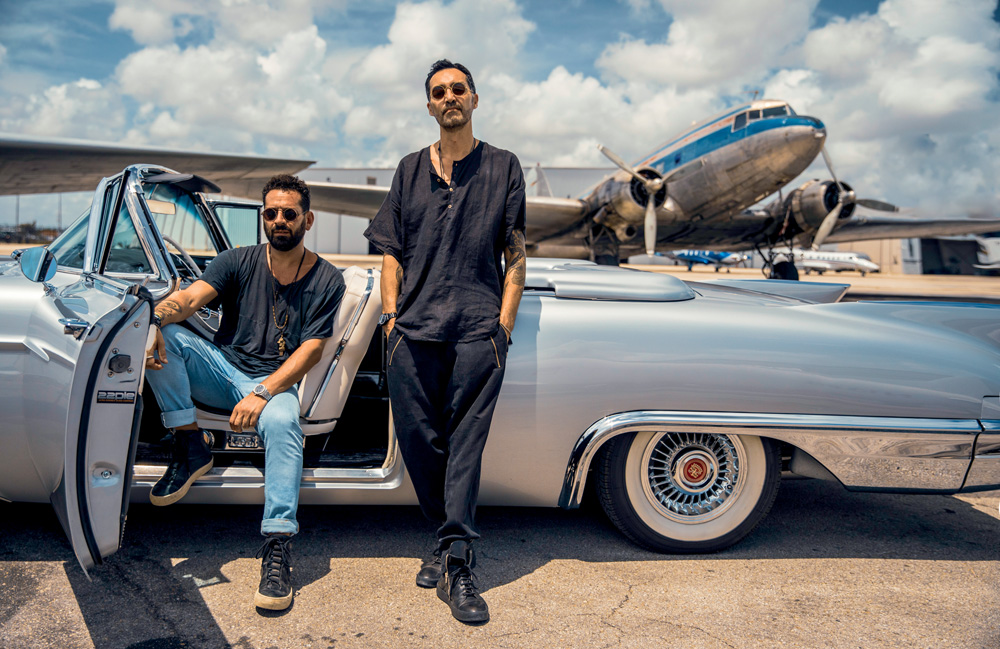 Duo Audiofly posa em carro conversível prata, ambos com camisas escuras. Um está sentado na poltrona do carro, com jeans claras, o outro de pé encostado.