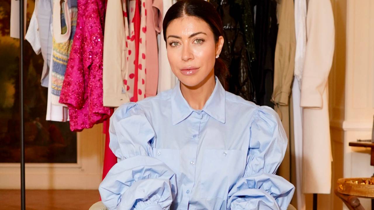 Carol Bassi, sentada de pernas cruzadas em poltrona branca, veste camisa azul claro com mangas bufantes e calça em tom de jeans escuro. Os cabelos castanho escuro estão presos em rabo de cavalo. Ao fundo, uma arara repleta de roupas.
