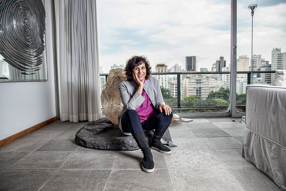 Imagem mostra mulher sentada em cristal, em sala com janela, ao fundo, aberta para um horizonte de prédios