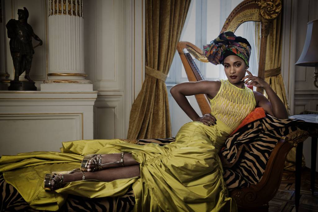ERIKA JANUZA (Fernando Torquatto-Divulgação) Moça negra posa sentada, quase deitada, em divã. Veste grande vestido na cor mostarda e tem uma das mãos na cintura.