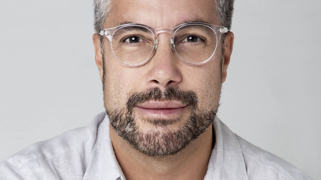 Fernando Torquatto encara a câmera sem sorrir. Veste camisa branca, tem cabelos grisalhos, barba preta e óculos de grau transparentes redondos.