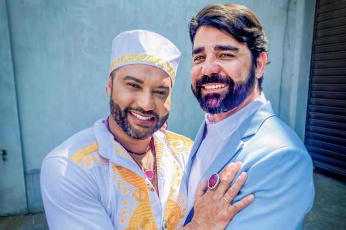 Rafael Celso Rodrigues e Allan Montenegro sorriem abraçados. Rafa veste turbante e roupa branca, Allan veste terno azul claro.