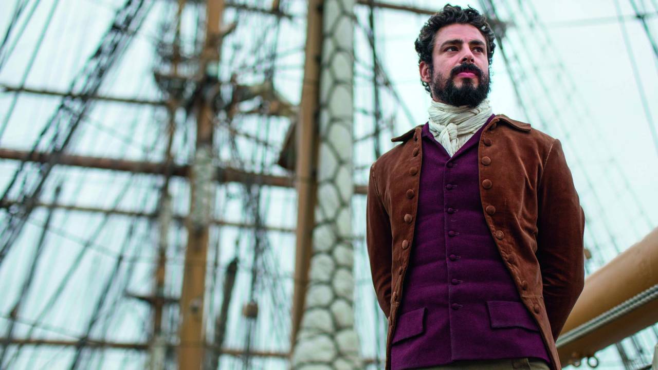 Imagem mostra homem de barba e roupa de gala com os braços para trás, em pé, com o mastro de uma caravela ao fundo