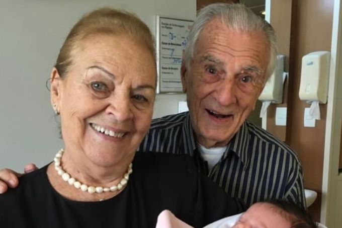 Glória Drummond e Orlando Drummond aparecem sorrindo. Ela segura uma bebê nos braços.