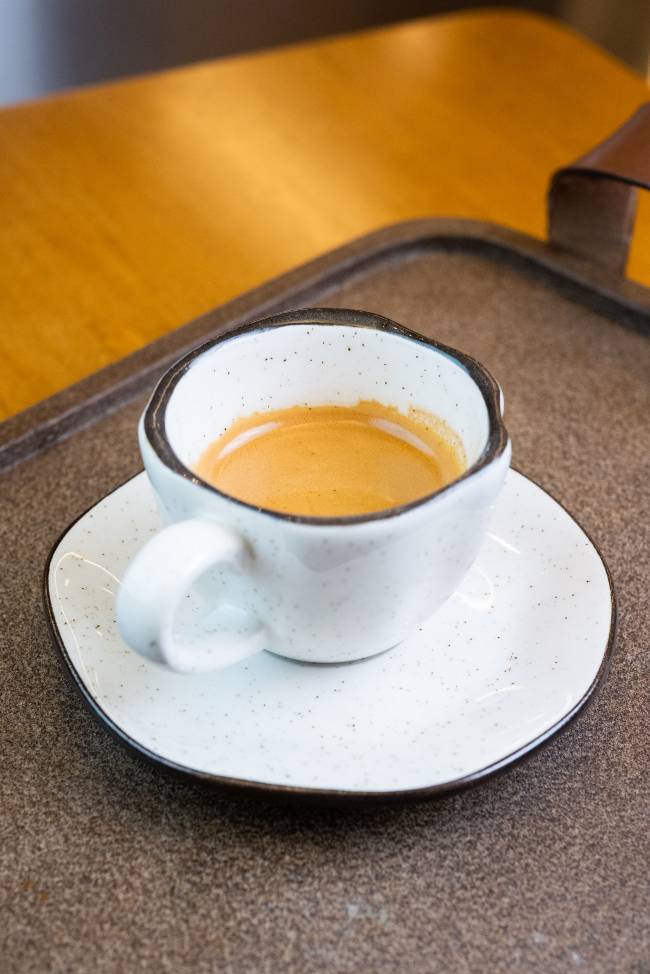 Café expresso baobá xícara branca com borda preta contendo café expresso sobre pires branco em uma bandeja marrom apoiada em uma superfície de madeira