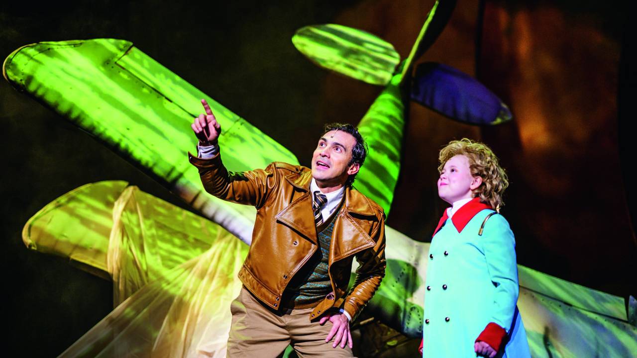 O personagem do Aviador aponta para cima e mostra algo para o Pequeno Príncipe no musical sobre o livro. No fundo, podemos ver uma tela de projeção com a imagem de uma avião verde