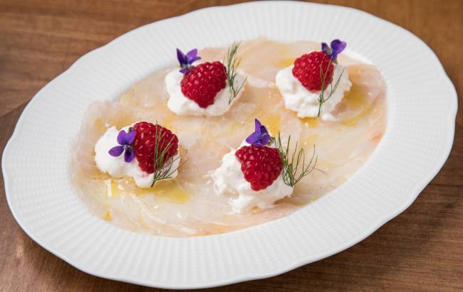 Prato branco com finas fatias de peixe cobrindo o fundo, quatro manchas de straciatella sobre as fatias, com uma framboesa e uma flor roxa em cada uma delas